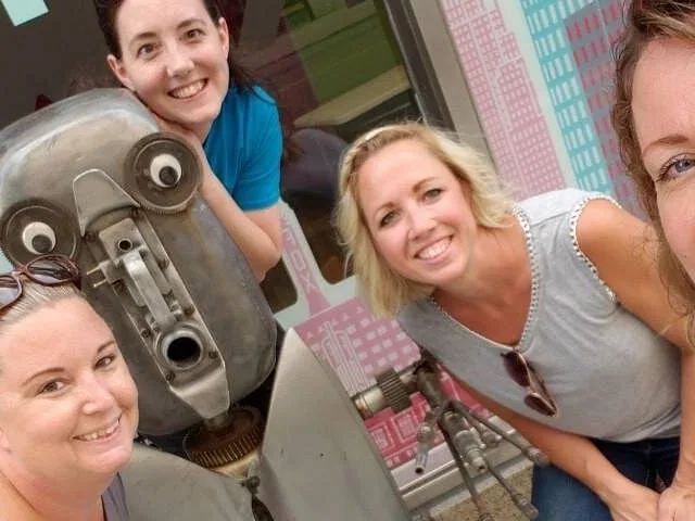 Gruppe von Frauen, die draußen mit einer mechanischen Skulptur lächeln